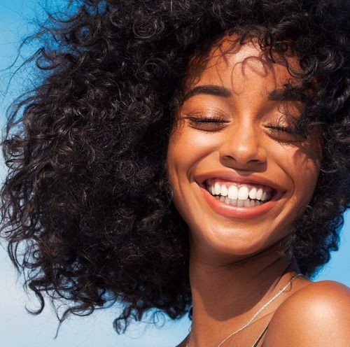 Woman with curly hair smiling broadly with eyes closed against a blue sky background.